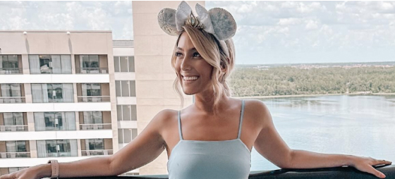 A guest wearing Minnie ears stands on a balcony overlooking a lake